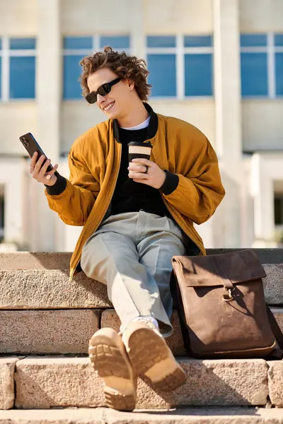 Um jovem na moda senta-se em degraus de pedra, tomando café e sorrindo para o telefone em plena luz do dia. — Fotografia de Stock