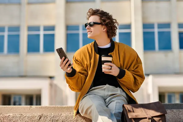 A stylish young man sits on a ledge, sipping coffee and smiling as he scrolls on his phone. — Stock Photo