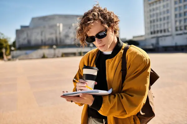 A young man with curly hair and sunglasses takes notes while enjoying coffee in a busy square. — Stock Photo