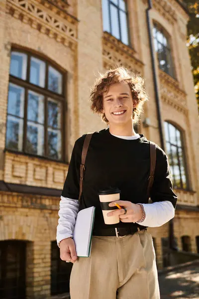 Charming young man holds a coffee cup and notebook while walking past historic campus architecture. — Stock Photo