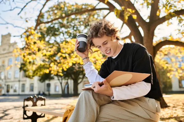 A young man sits on a bench in a sunlit park, reading and enjoying his coffee amid autumn leaves. — Stock Photo