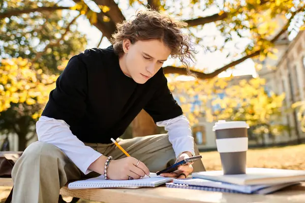 A young man carefully writes notes and checks his phone, surrounded by vibrant autumn leaves. — Stock Photo