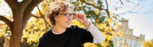 A young man with curly hair and glasses smiles while basking in the sunlight next to a tree. — Stock Photo