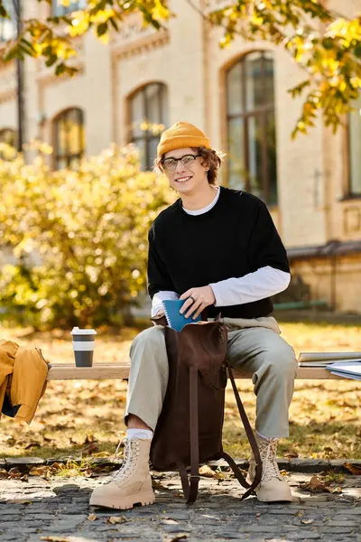 A young man with a fashionable look enjoys a warm drink while resting on a bench in a park. — Stock Photo