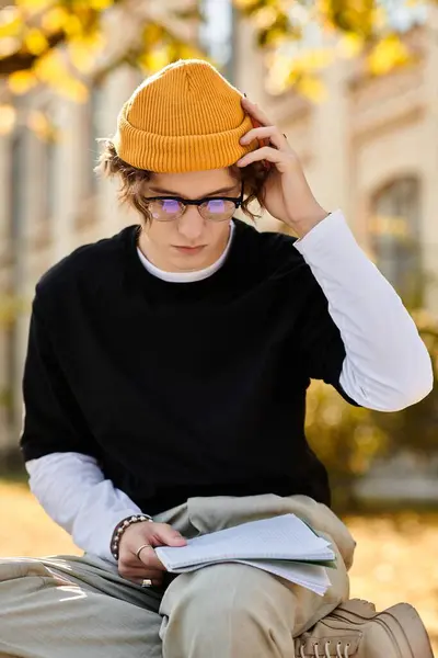 A young man in a yellow beanie focuses intently on his notes while sitting in a sunny park. — Stock Photo