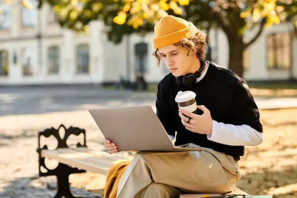 Sitting on a bench, a young man focuses on his laptop, enjoying a warm cup of coffee in autumn. — Stock Photo