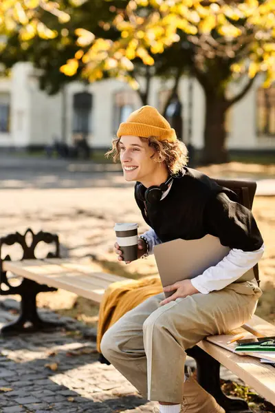 A young man sits on a park bench with coffee, studying under autumn leaves. — Stock Photo
