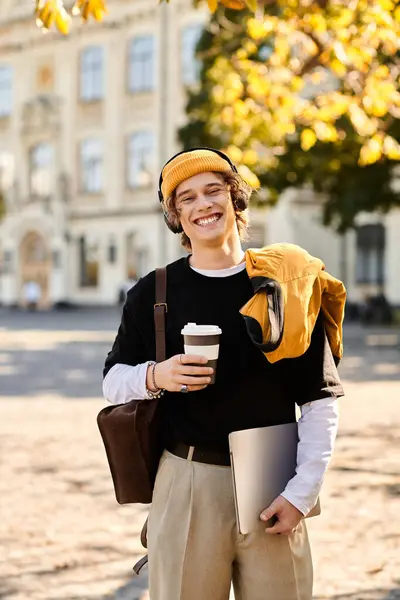 A young man smiles with a coffee and laptop on a university campus amid autumn leaves. — Stock Photo