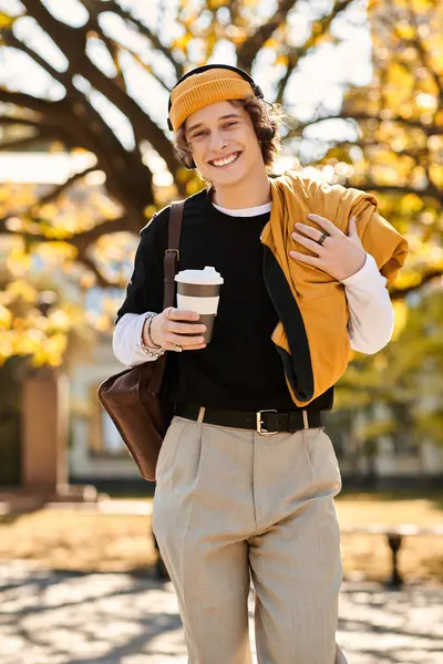 A young man with a stylish look strolls through a park, sipping coffee while wearing autumn attire. — Stock Photo