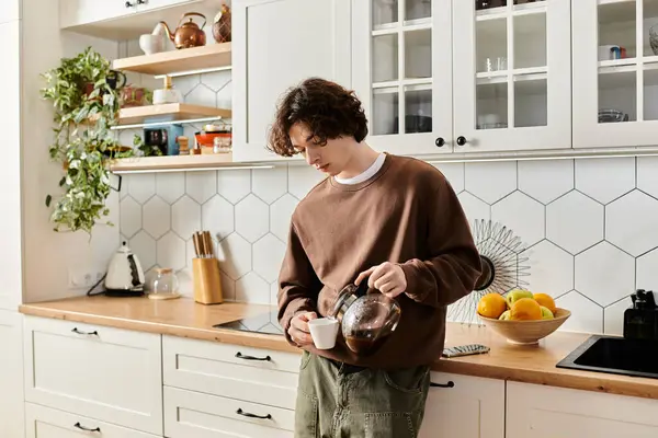 Un joven con una sudadera marrón vierte su café de la mañana en una cocina elegante llena de luz. - foto de stock