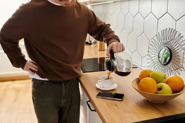 Curly-haired student in a brown sweatshirt enjoys coffee preparation in his modern apartment. — Stock Photo