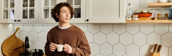 A good looking young man with curly hair stands in his stylish kitchen savoring coffee, banner — Stock Photo