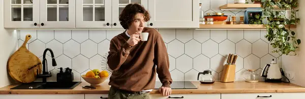A young man in a brown sweatshirt sips a drink while relaxing in his stylish kitchen at home, banner — Stock Photo