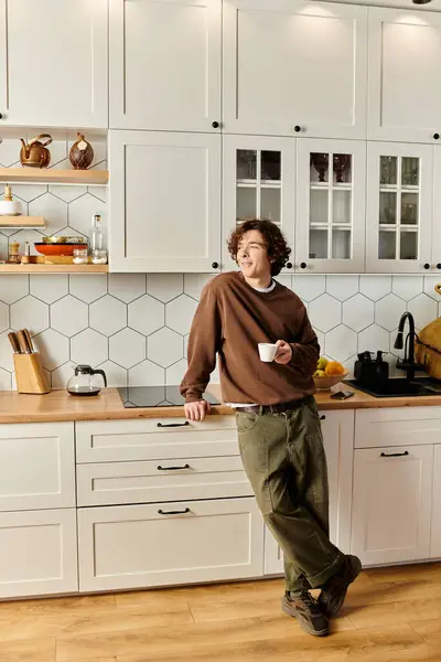 Curly-haired young man relaxes with a cup in his modern kitchen amid warm decor. — Stock Photo