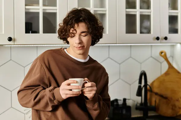 Young man in a brown sweatshirt savors his coffee, reflecting in a modern kitchen setting. — Stock Photo