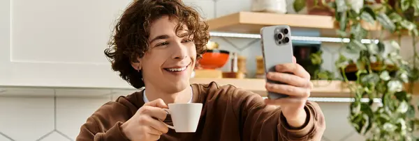 In a contemporary apartment, a young student with curly hair sips coffee and smiles at his phone, banner — Stock Photo