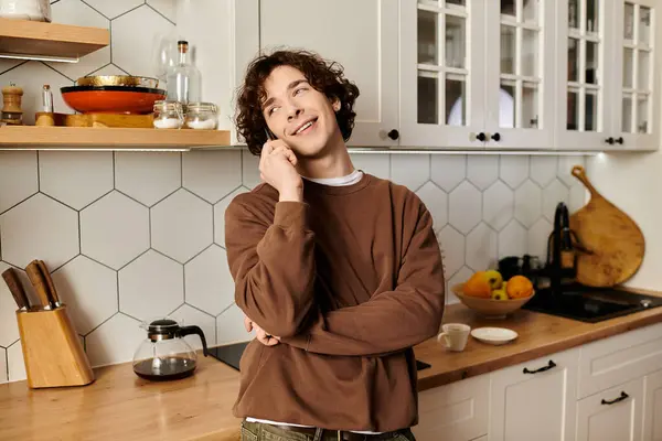 Joven estudiante con el pelo rizado disfruta de una animada llamada telefónica en su elegante espacio de cocina. - foto de stock