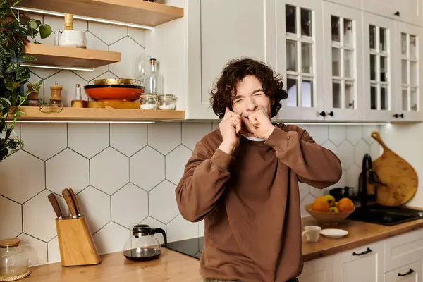 A young man in a brown sweatshirt playfully stretches his face in a stylish kitchen. — Stock Photo