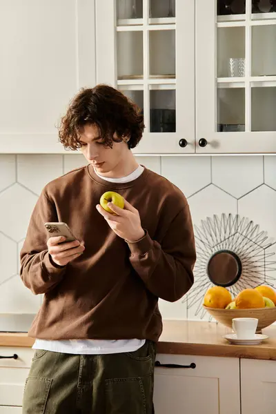 Jovem em uma camisola marrom relaxa em casa, navegando em seu telefone com uma maçã na mão. — Fotografia de Stock