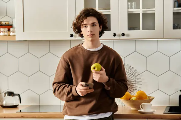 A young man in a brown sweatshirt holds an apple and a phone, showcasing his cozy apartment. — Stock Photo