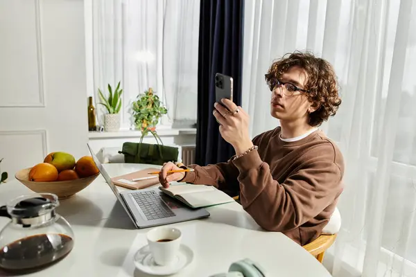 Young man in a brown sweatshirt interacts with his smartphone at a stylish table while studying. — Stock Photo
