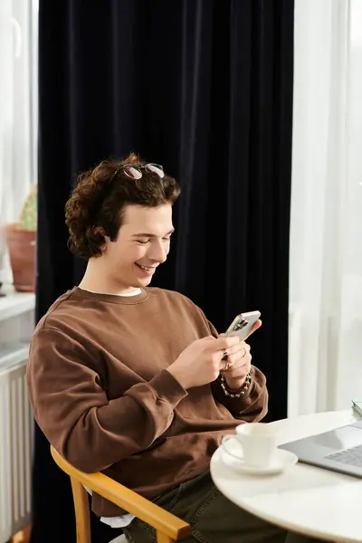 Young man in brown sweatshirt smiles while using his smartphone in a cozy urban apartment. — Stock Photo