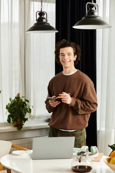 Un giovane uomo con i capelli ricci sorride in un appartamento moderno, con in mano un telefono e un portatile. — Foto stock