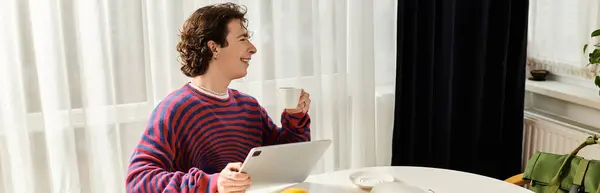 A non binary individual in a striped sweatshirt smiles while sipping coffee at home, banner — Stock Photo