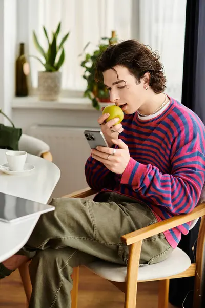 In a stylish apartment, a young non-binary person munches an apple while on their phone. — Stock Photo