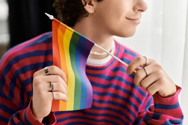 Un giovane individuo non binario sorride mentre tiene una bandiera orgoglio nel loro spazio accogliente e contemporaneo. — Foto stock
