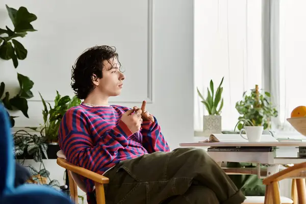 A young non-binary individual relaxes in a colorful sweatshirt while surrounded by plants. — Stock Photo