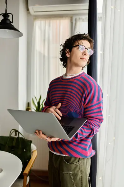 In a bright modern apartment, a young non binary person holds a laptop, contemplating ideas. — Stock Photo