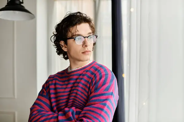 A young non-binary person with curly hair reflects while gazing out a window in a modern apartment. — Stock Photo