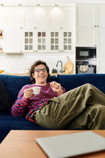 A young non binary individual relaxes on a couch, sipping coffee and embracing a cozy moment. — Stock Photo