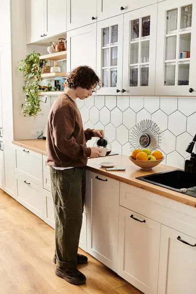 Joven con una sudadera marrón vierte una bebida en su elegante cocina llena de luz natural. - foto de stock