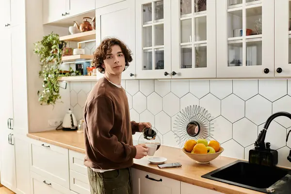 A young student in a brown sweatshirt pours coffee in a modern kitchen filled with natural light — Stock Photo