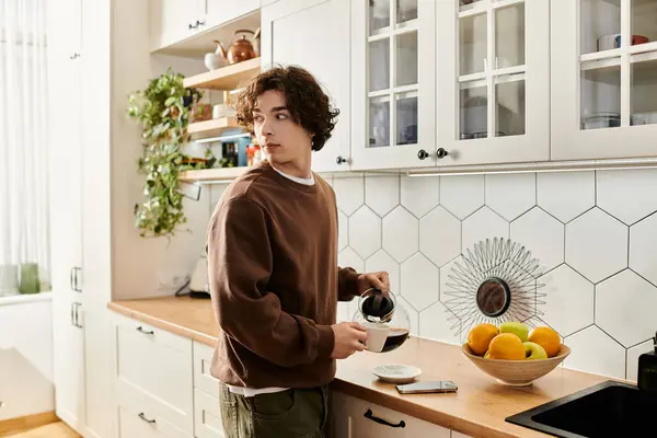 Joven con una sudadera marrón vierte café en su elegante cocina, saboreando el momento. - foto de stock