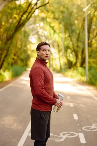 Handsome man with tattoos relaxes in a green park, savoring a drink amidst natures beauty. — Stock Photo