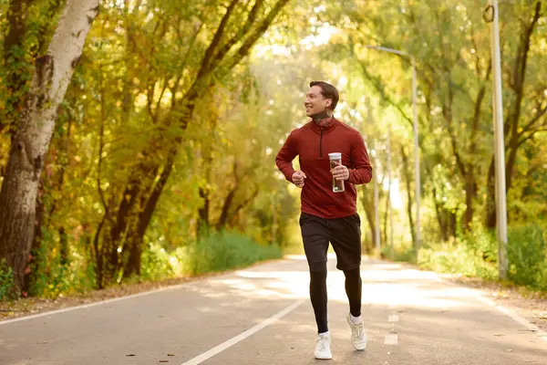 Handsome man in a trendy outfit jogs along a picturesque pathway surrounded by vibrant trees. — Stock Photo