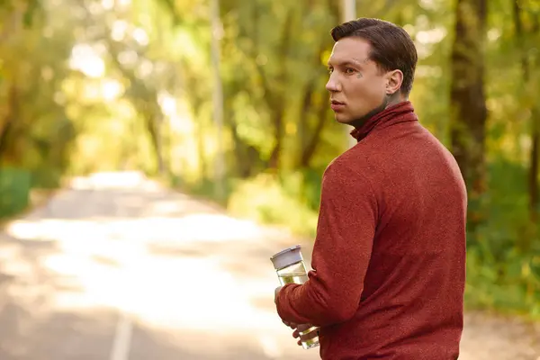 Handsome man in modern attire poses outdoors, holding a beverage while exploring nature. — Stock Photo