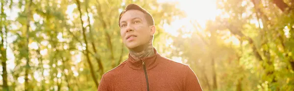 Un joven tatuado se encuentra confiado en un bosque iluminado por el sol, ostentando un atuendo moderno. - foto de stock