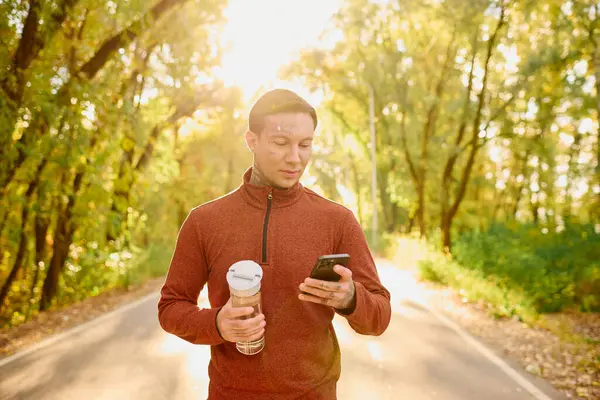 A young man with tattoos strolls a scenic path, drink in hand, checking his phone. — Stock Photo