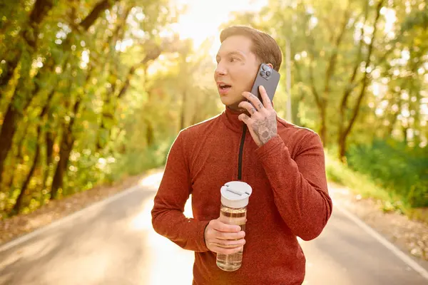 Handsome man with tattoos holds a water bottle while talking on the phone in a lush green setting. — Stock Photo