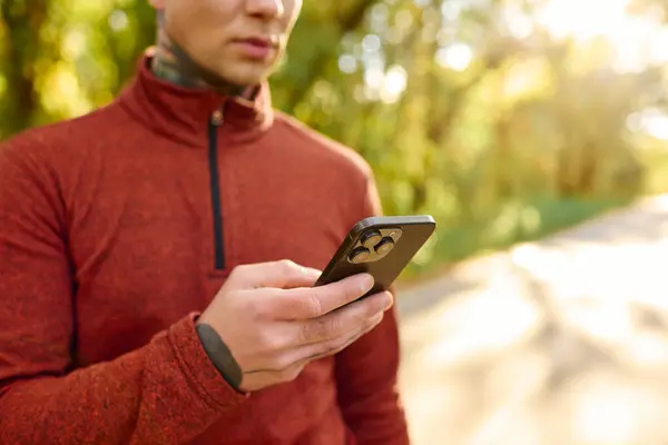 A stylish young man with tattoos stands outdoors, focused on his smartphone in the sun. — Stock Photo