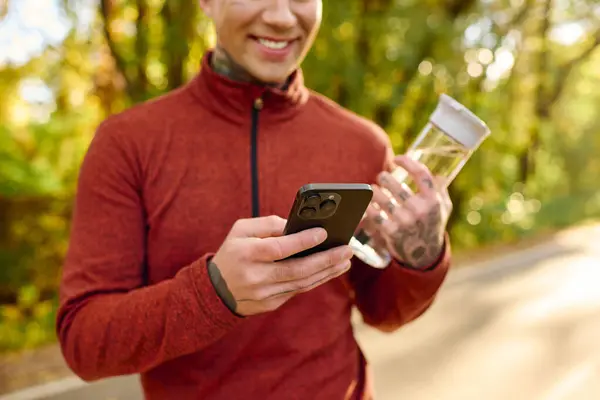 Handsome man with tattoos checks his smartphone while holding a water bottle in a vibrant park. — Stock Photo