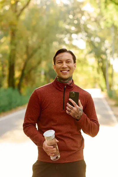 A young man with tattoos smiles while holding a water bottle, surrounded by colorful autumn trees. — Stock Photo