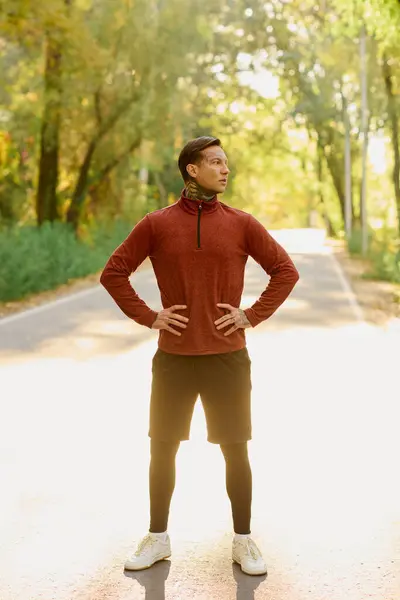 Handsome man with tattoos standing proudly on a sunlit path surrounded by trees in autumn. — Stock Photo