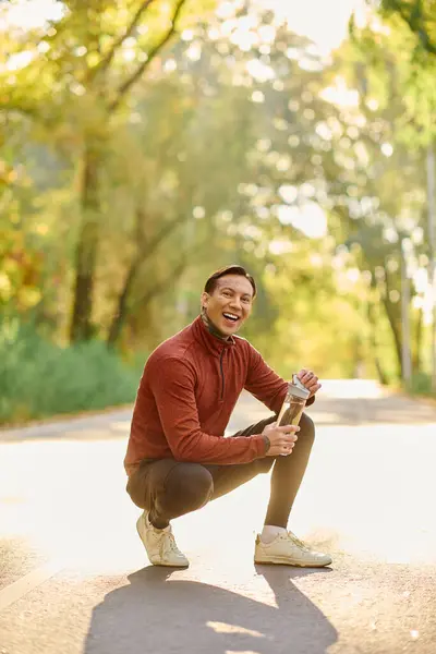 Handsome young man kneels on a paved path, savoring a drink surrounded by autumn foliage. — Stock Photo