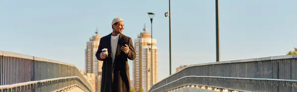 Schöner Herr in trendiger Kleidung geht selbstbewusst auf eine Brücke, nippt am Kaffee und posiert. — Stockfoto