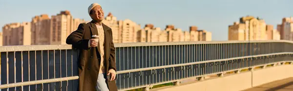 Un hombre tatuado con estilo disfruta de una bebida en un puente, viendo la puesta de sol sobre la vida de la ciudad. - foto de stock